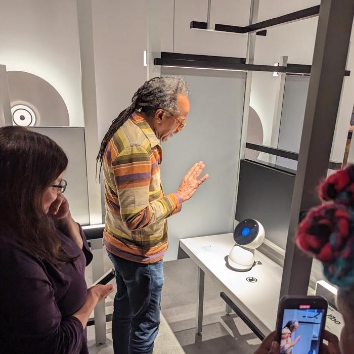 Fellow Louis Massiah waves to Jibo, a small social robot sitting on a table.